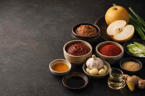 Korean pantry staples on a dark surface: bowls of doenjang, gochujang, and gochugaru with Asian pear, scallions, garlic, ginger, sesame seeds, oil, and soy sauce.